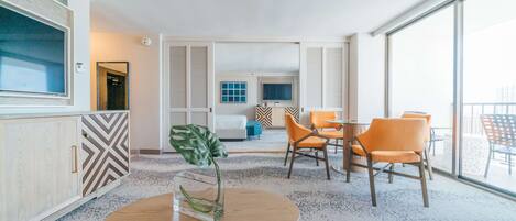 A modern hotel room with a large window, a round wooden table with a vase, two orange chairs, a bed, and a TV.