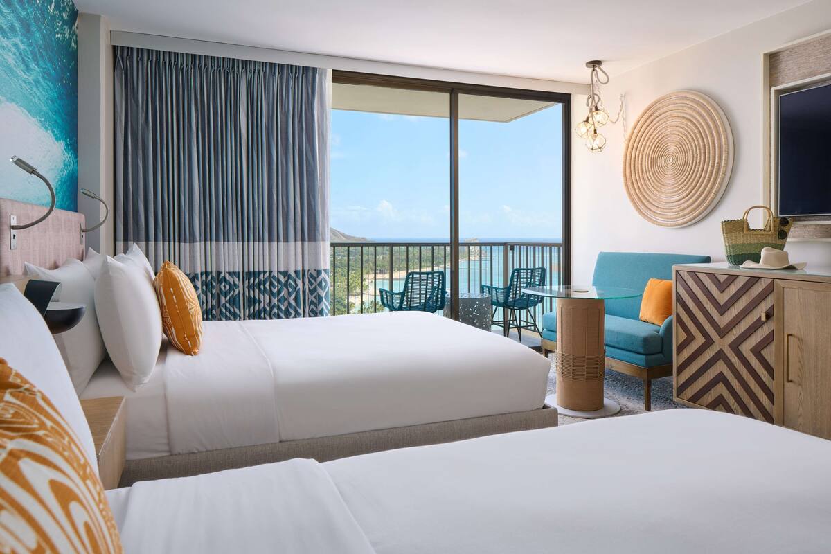 A hotel room with two beds, a balcony with a view, a TV, and a seating area with a table and chairs.