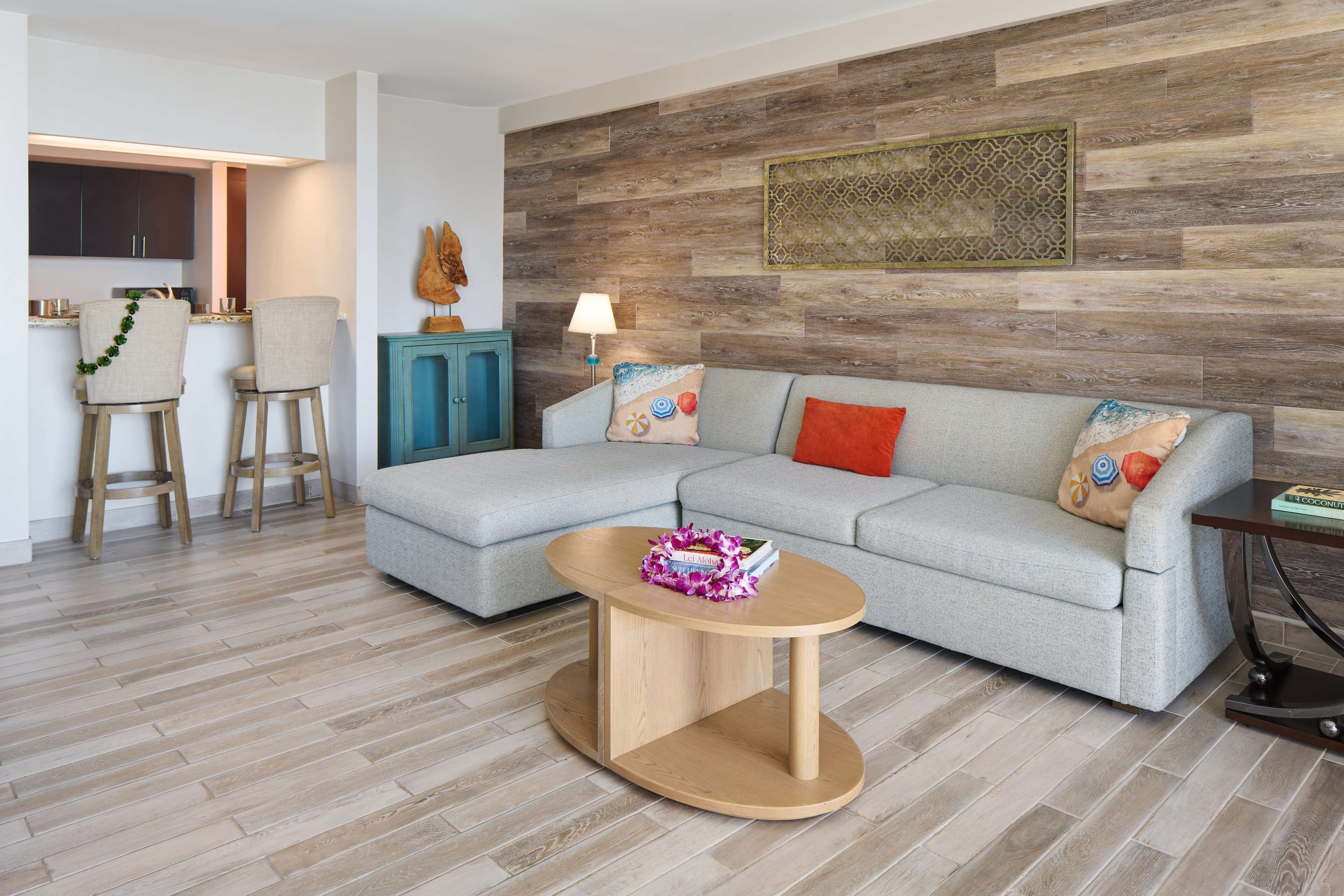 A modern living room with a grey sectional sofa, a wooden coffee table, and a kitchen area with dark cabinets and a blue cabinet.