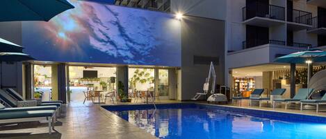 Una piscina interior con sillas de descanso y sombrillas, una pantalla digital grande que muestra una imagen del cielo, y un edificio moderno con balcones.