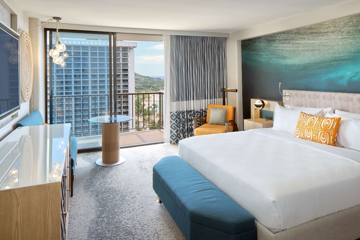 A hotel room with a large bed, a desk, a chair, a balcony with a view of buildings and mountains, and a large window with curtains.