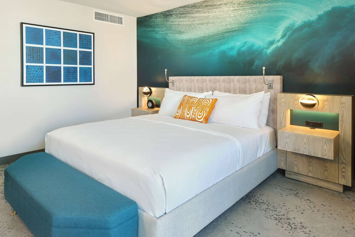 A modern hotel room with a large bed, a bench, a wall mural, and a framed picture.