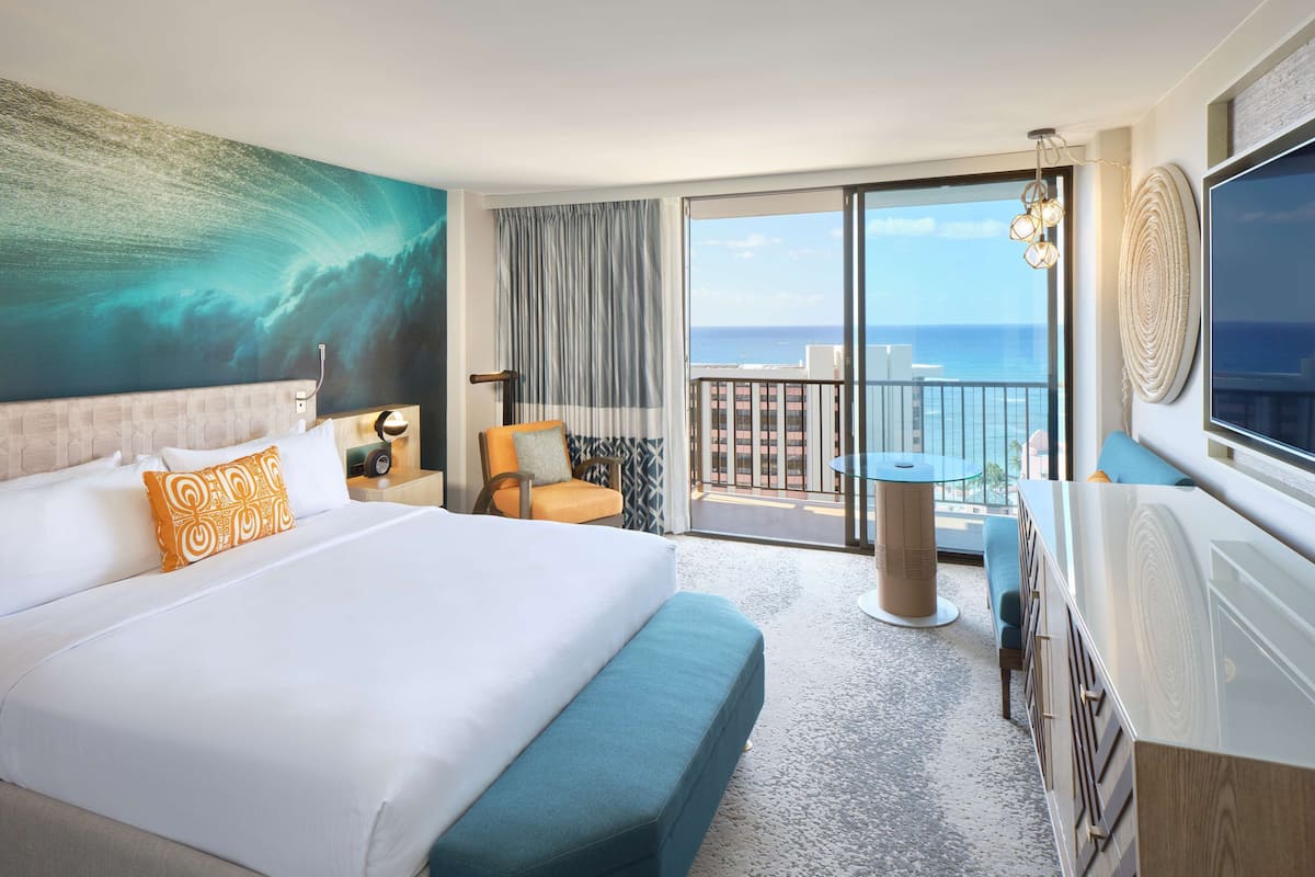 A hotel room with a large bed, a chair, a small table, a TV, and a balcony with a view of the ocean.
