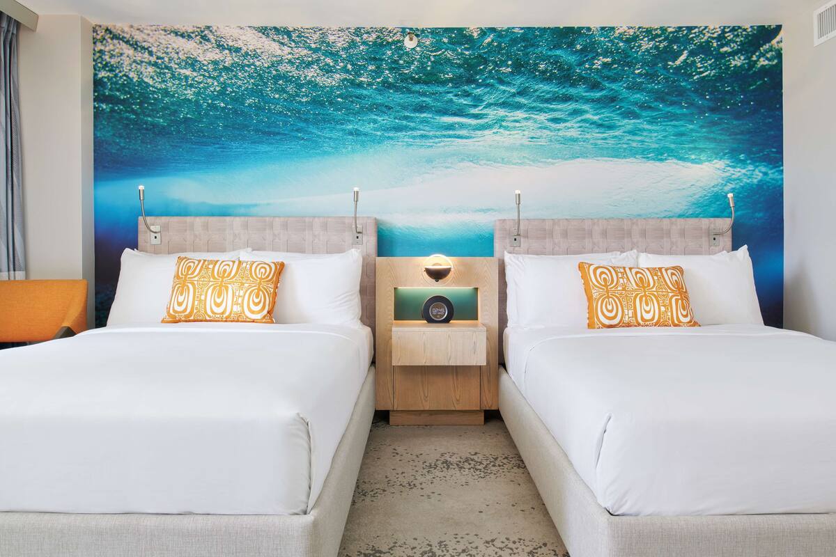 A hotel room with two beds, a nightstand with a lamp, and a large ocean-themed wall mural.