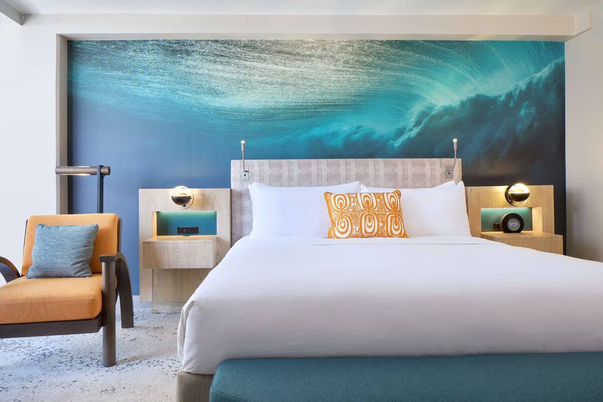 A hotel room with a large bed, a nightstand with a lamp, a chair, and a wall mural of an underwater scene.