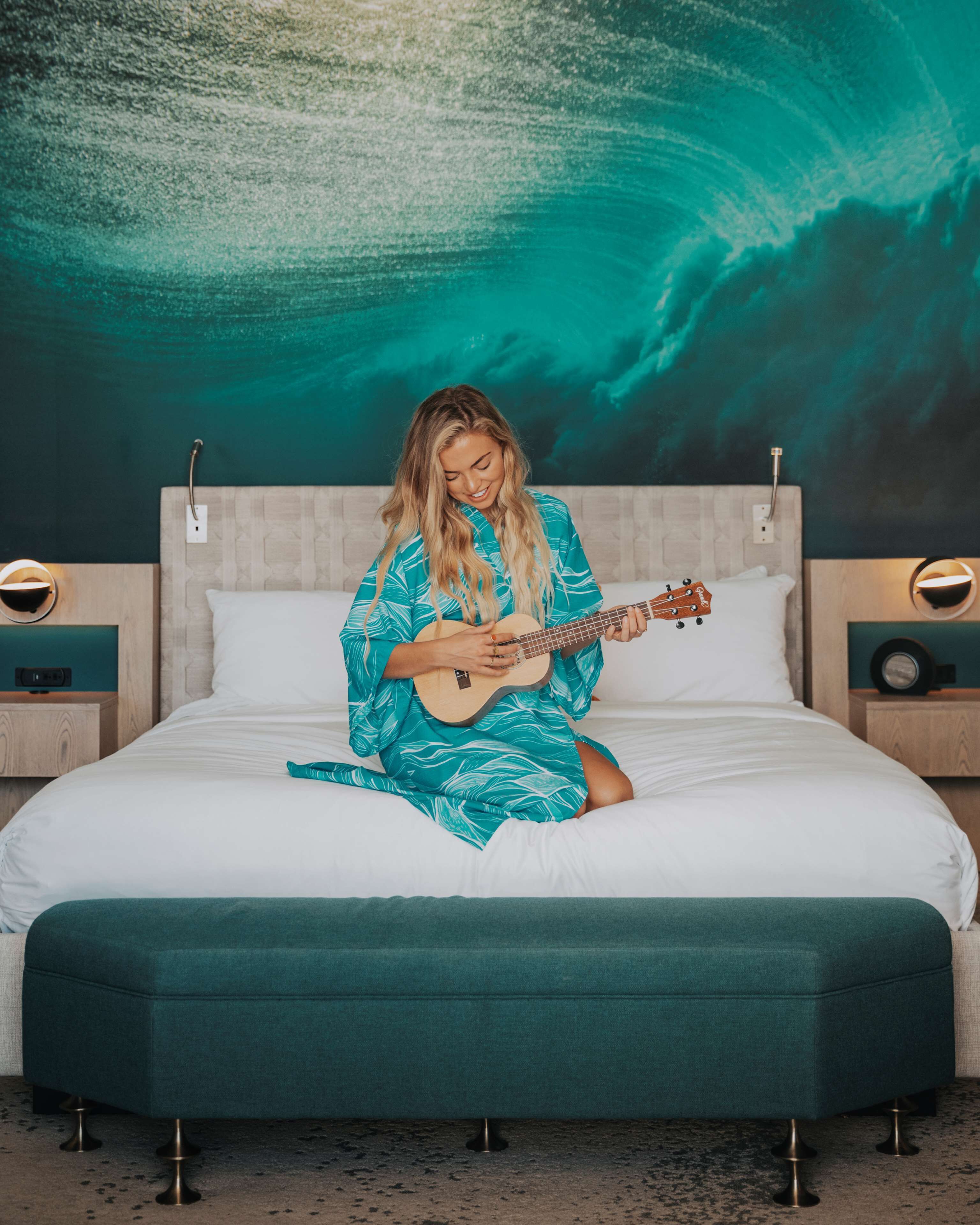 A person sitting on a bed with a guitar, a large bedspread, and a wall mural.