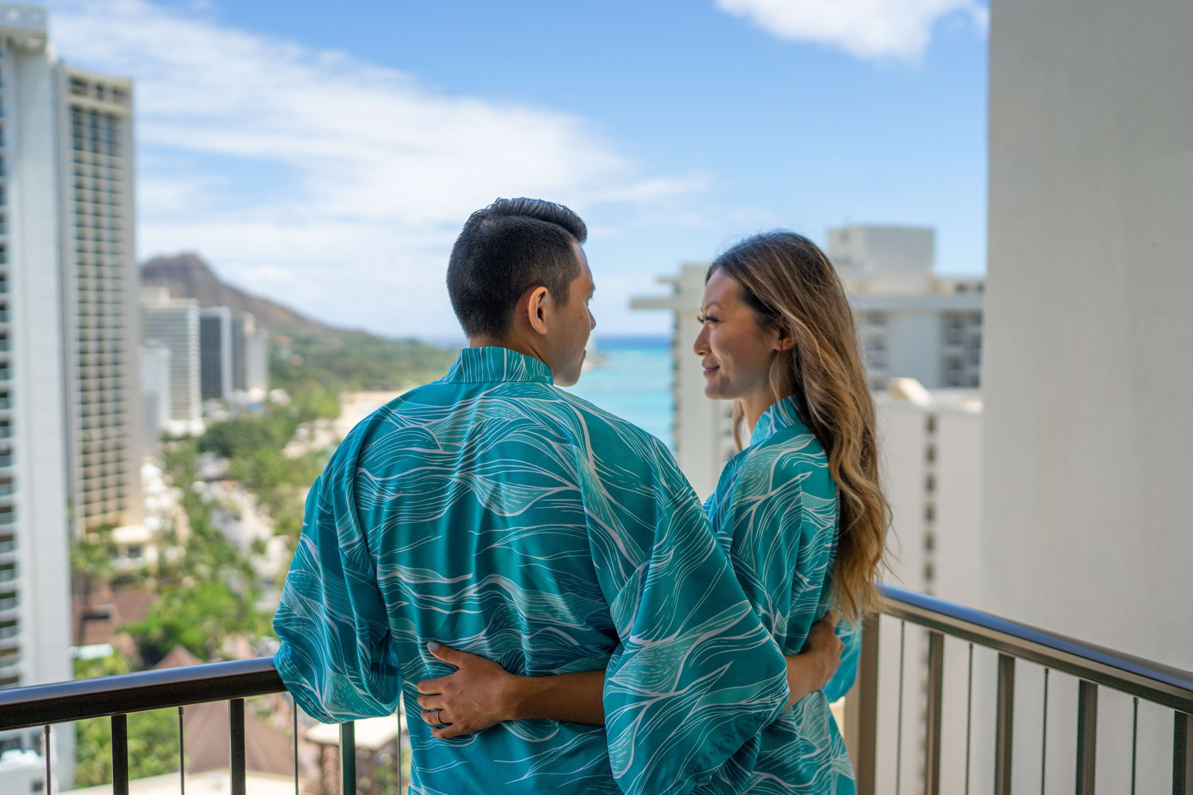 A couple standing on a balcony overlooking a cityscape with buildings and a distant mountain.