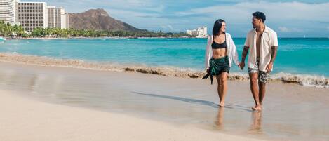 A couple walking on a beach with a cityscape in the background.