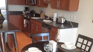 A dining area with a dark wooden table set for four, surrounded by matching chairs. A kitchen with wooden cabinets and stainless steel appliances is visible in the background. - Weichert Suites at Arlington Courthouse (Arlington)