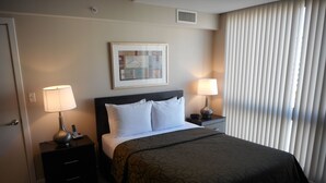A hotel room with a bed, two bedside tables with lamps, a nightstand, a wall-mounted artwork, and a window with blinds. - Weichert Suites at Arlington Courthouse (Arlington)