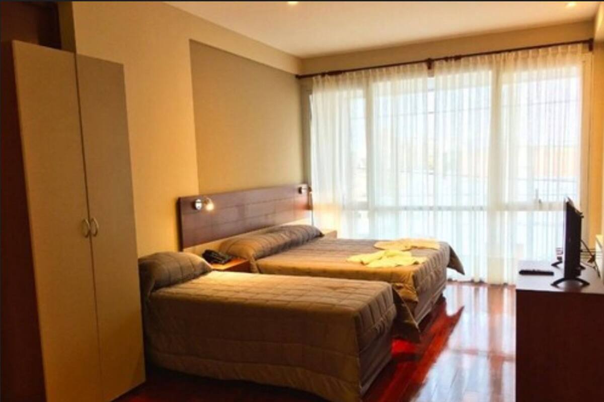 A hotel room with two beds, a wardrobe, a desk with a computer, and a large window with sheer curtains.