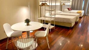 Une chambre d’hôtel avec une table à manger ronde, deux chaises blanches, un porte-vêtements suspendu et deux lits avec du linge de lit blanc.