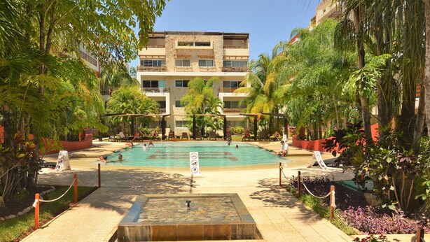 A pool area with a stone fountain, surrounded by palm trees and lounge chairs. - Condo Sabia Apartments by CSR (Playa del Carmen)