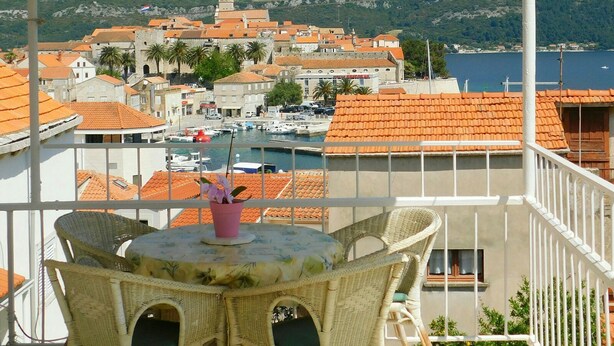 A balcony with wicker furniture overlooking a coastal town with red-roofed buildings and a marina. - Apartments Neno (Korcula)