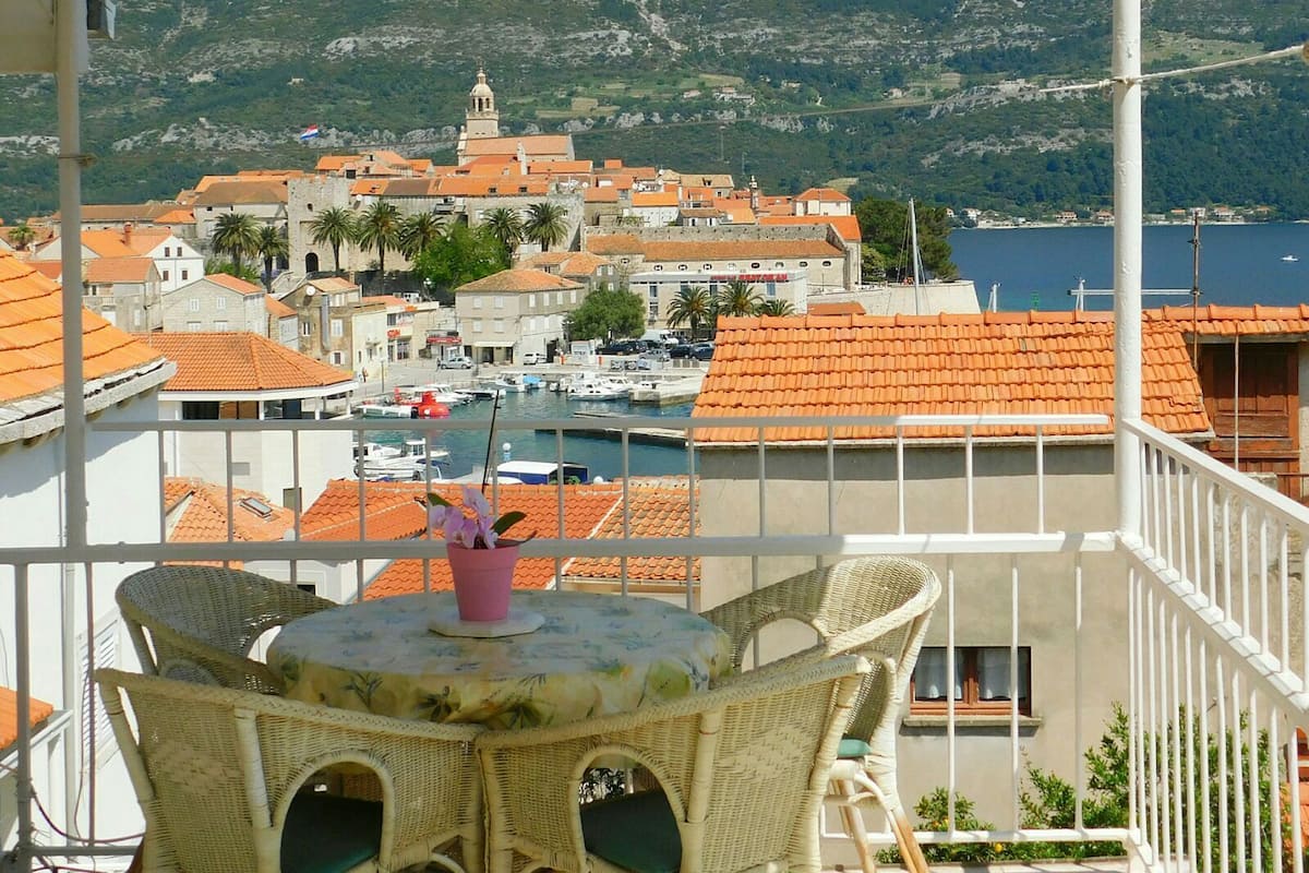 Un balcón con mobiliario de mimbre con vistas a un pueblo costero de edificios con tejados rojos y un puerto deportivo.