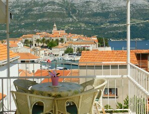 A balcony with wicker furniture overlooking a coastal town with red-roofed buildings and a marina. - Apartments Neno (Korcula)