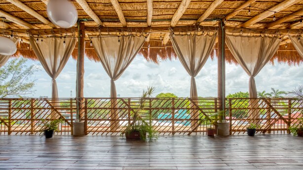 Una terraza de madera abierta con techo de paja, plantas en macetas y vistas a una piscina.