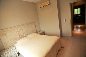 A bedroom with a bed, a nightstand, an air conditioning unit, and a door leading to another room.