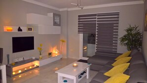 Een moderne woonkamer met een flatscreen televisie, een witte salontafel, een grijze bank met gele kussens, en een groot raam met zonwering.
