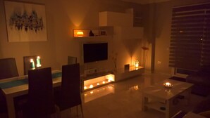 Een gedimd verlichte woonkamer met een flatscreen televisie, een bijzettafeltje en verschillende kaarsen.