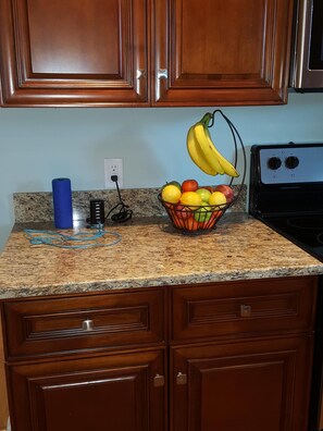 Une cuisine avec des meubles de rangement en bois, un plan de travail en granit, un panier de fruits, un objet cylindrique bleu et une cuisinière.