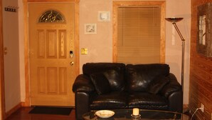 Una sala de estar con una puerta de madera, un sofá de cuero negro, una mesa de centro de vidrio y una lámpara de pie.