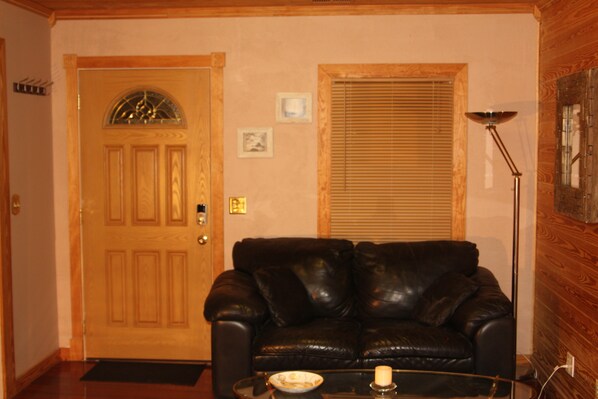A living room with a wooden door, a black leather sofa, a glass coffee table, and a floor lamp.