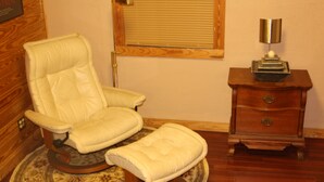 Una habitación con un sillón de cuero y reposapiés sobre una alfombra estampada, una mesita de madera con una lámpara y suelo de madera.