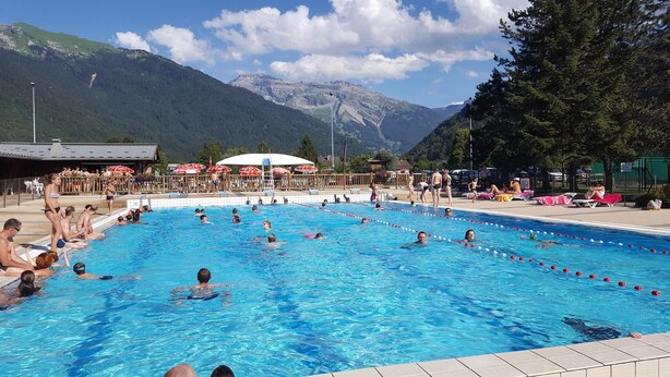 Piscina com pessoas nadando e tomando sol, rodeada por montanhas e guarda-sóis.