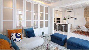 Ein modernes Wohnzimmer mit einer Couch, Hocker und einem Glastisch. Es gibt eine Küchenzeile mit einer Bar und Sitzgelegenheiten.