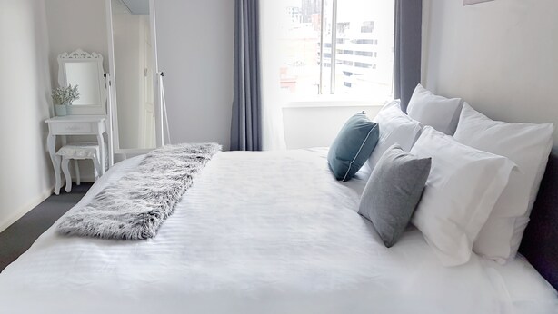 A modern bedroom with a large bed, a small desk, a mirror, and a view of the city through the window. - King Deluxe Apartments (Melbourne)
