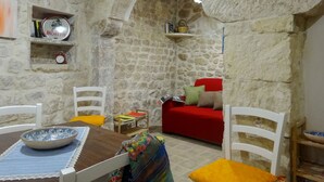 A stone-walled room with a red sofa, a wooden table with a bowl, and white chairs.