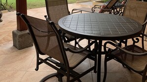 Un patio cubierto con una mesa redonda y sillas de mimbre, con vista a un área verde y el océano.