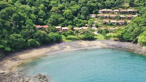 Vista aérea de un resort con varias edificaciones rodeadas de un bosque frondoso, una playa de arena y aguas turquesas cristalinas.