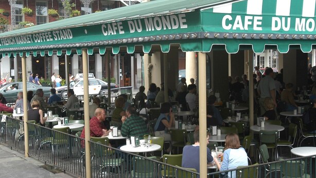 Café al aire libre con mesas y sillas, personas comiendo, y un toldo verde con señalización.
