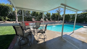 En overdækket terrasse med pool, udendørs møbler og udsigt over poolen.