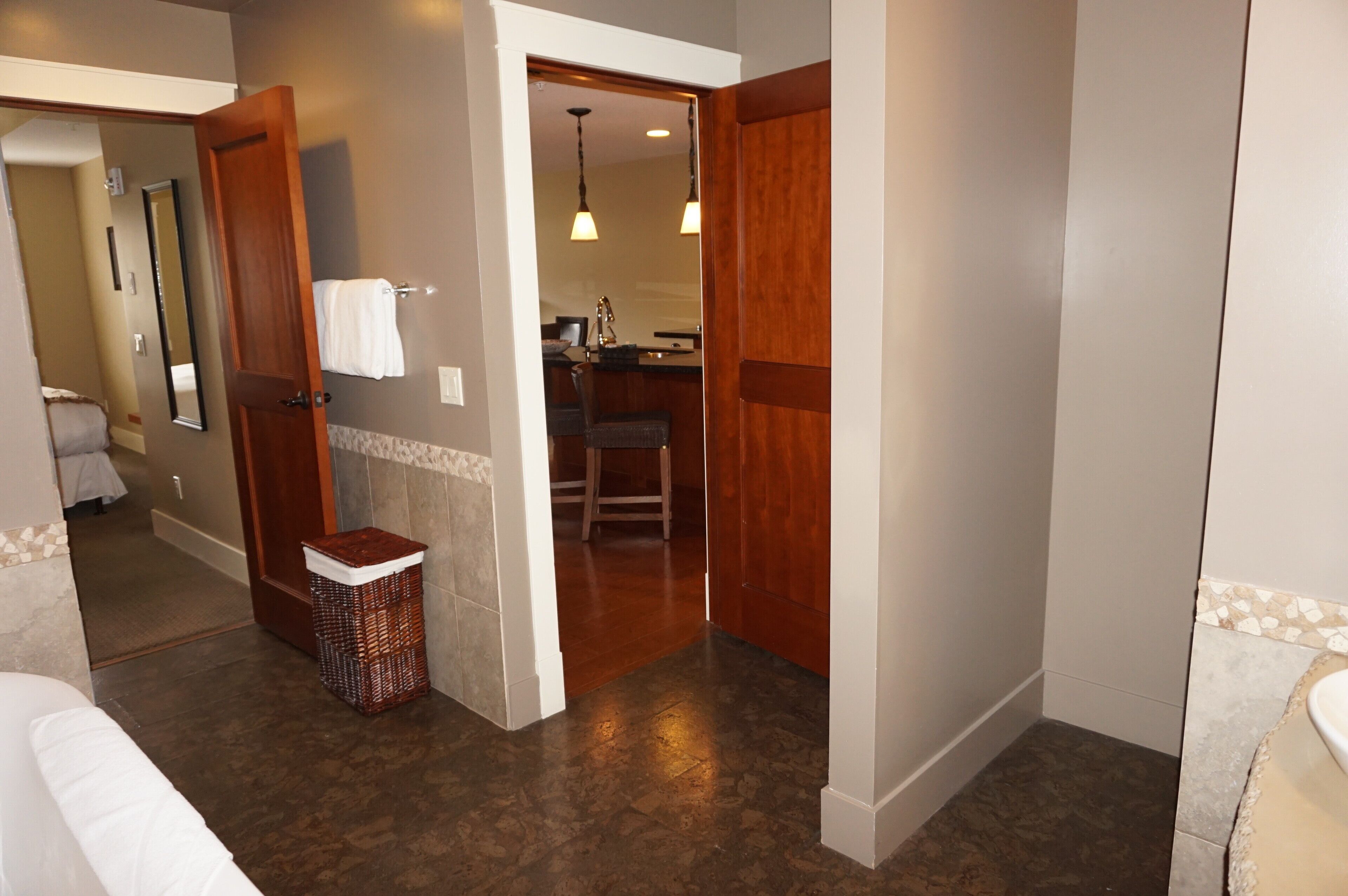 A hotel room with a wooden door, a small wicker basket, a white towel on a hook, and a visible bathroom area with a sink and mirror.
