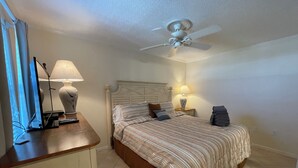 A bedroom with a bed, bedside lamps, a ceiling fan, and a desk with a lamp and a computer monitor.