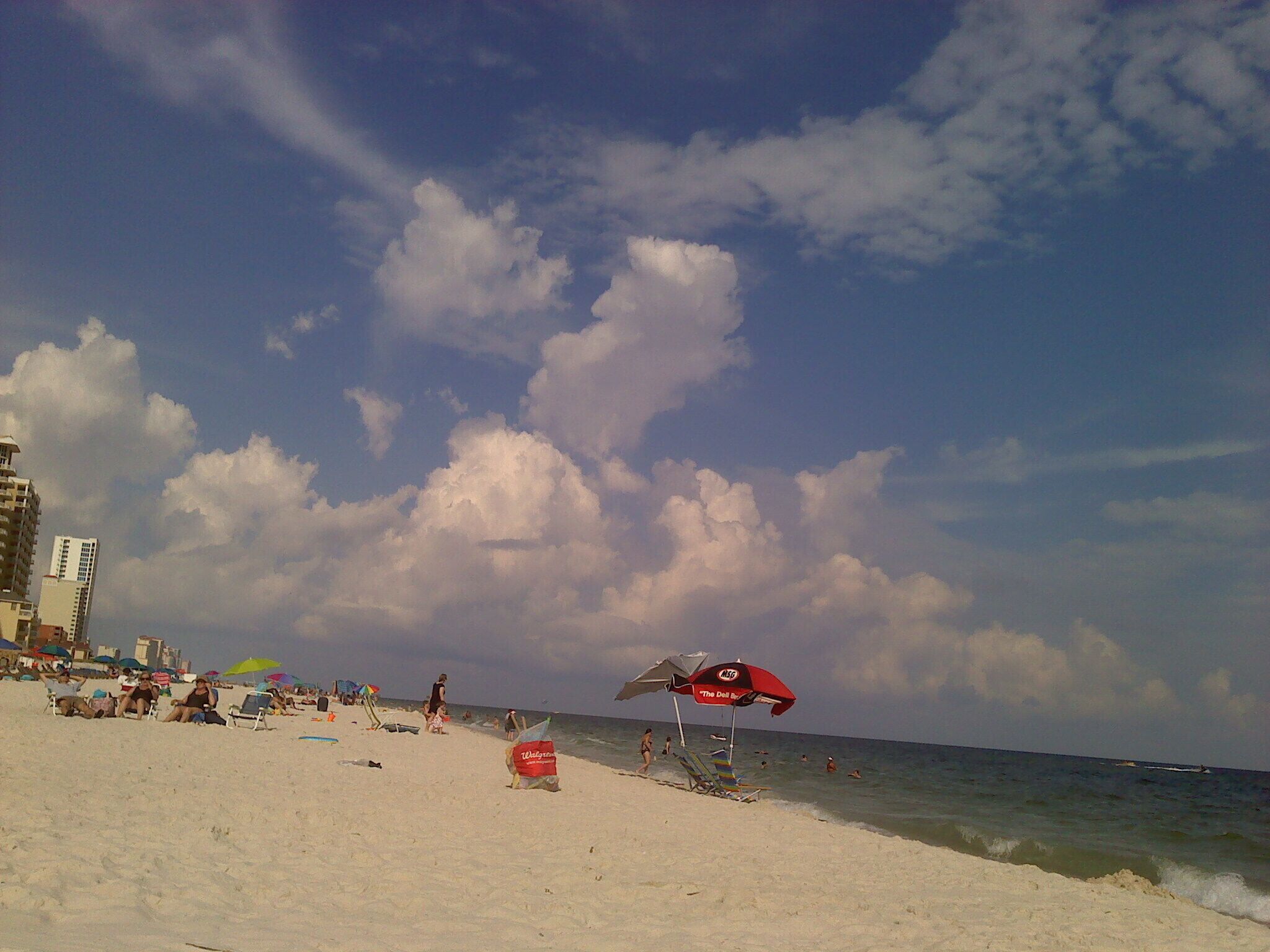 A beach with people sunbathing and swimming, umbrellas, and buildings in the background.