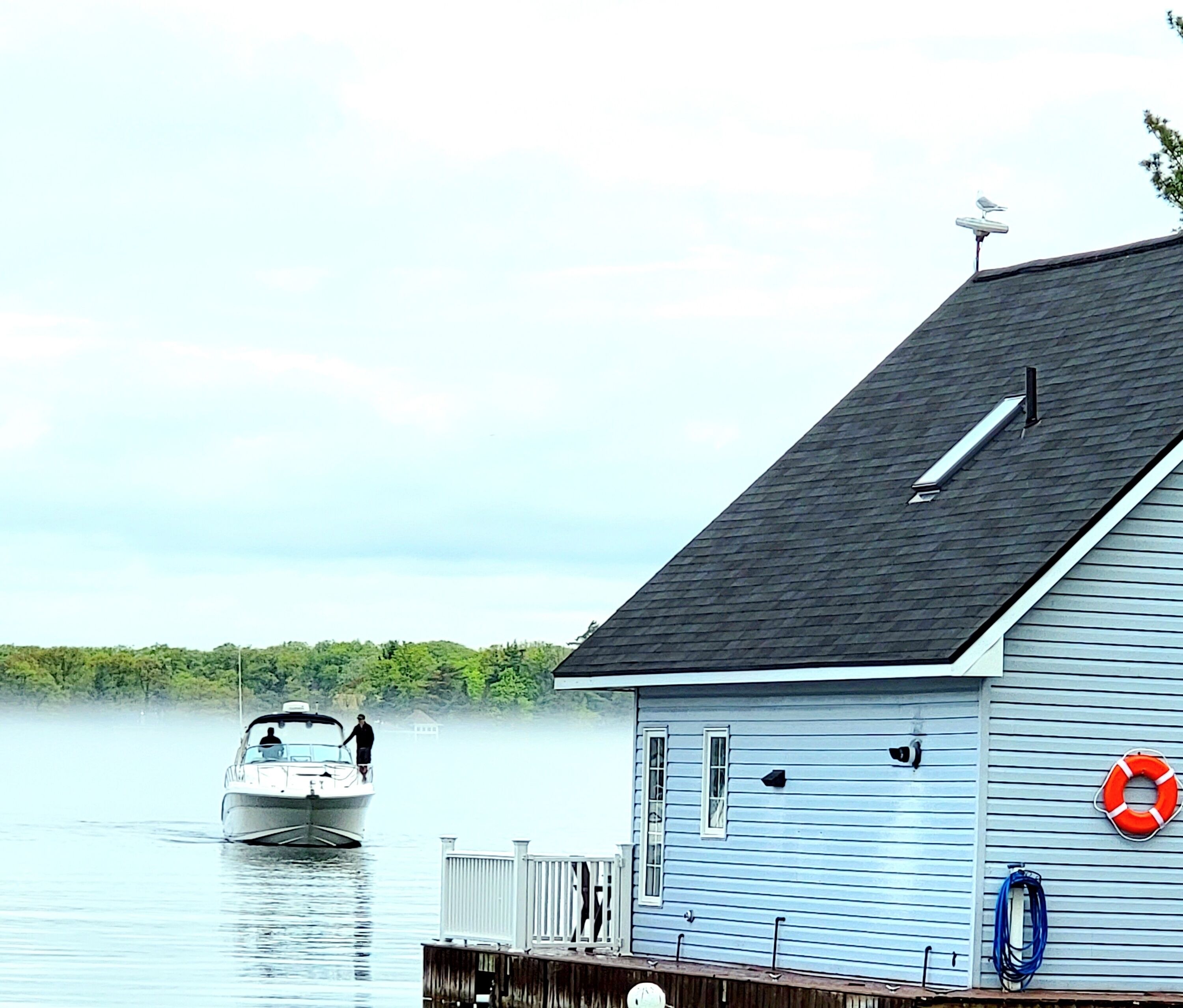 A house with a dock extending into calm water, a boat on the water, and a life preserver on the dock.