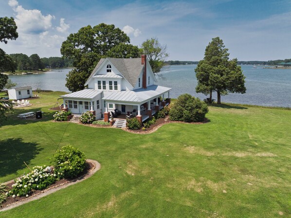 A large white house with a porch, surrounded by a well-maintained lawn and a lake view. - Experience the Best of Mathews Riverfront Living here at Mariner's Legacy! (Bohannon)
