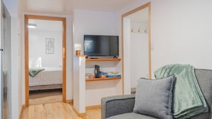 Een kleine, goed verlichte kamer met een bed, een televisie op een plank, een grijze bank en een spiegel.