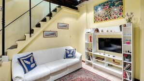 A living room with a white sofa, a TV on a white stand, a staircase, and a painting on the wall. - PF Apartment 2 Bedroom- and you're in Piazza Navona CIN:IT058091C2SLMC5MBC (Roma)