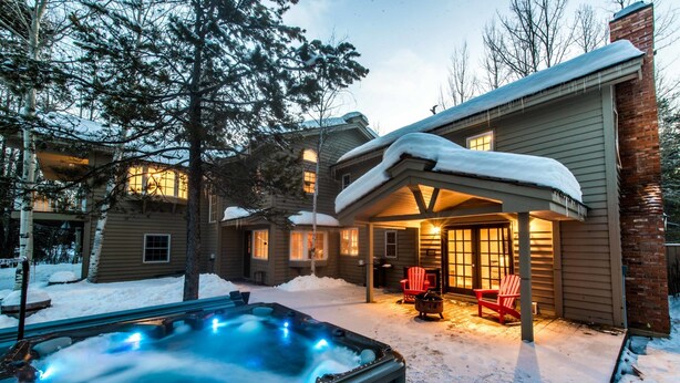 A snow-covered outdoor area with a hot tub, red Adirondack chairs, and a wooden house with multiple windows.