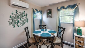 Un comedor con una mesa redonda, cuatro sillas, un equipo de aire acondicionado, decoración en la pared y una ventana con cortinas.