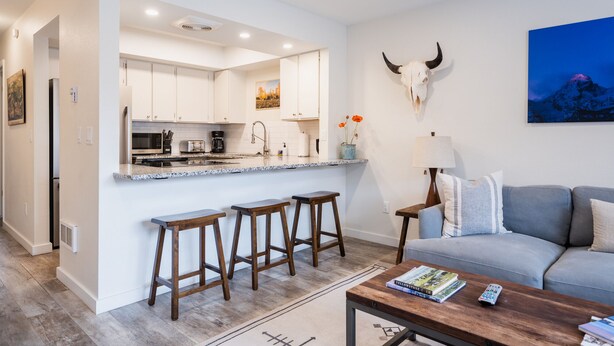 Una cocina moderna con armarios blancos, encimera de granito y una zona de comedor con taburetes. Hay una sala de estar con sofá, mesa de centro y un cráneo de animal montado.