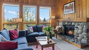 A cozy living room with a stone fireplace, a blue sofa, and a large window offering a scenic view. - Outpost: Nez Perce D5- Hot Tub Access (Teton Village)