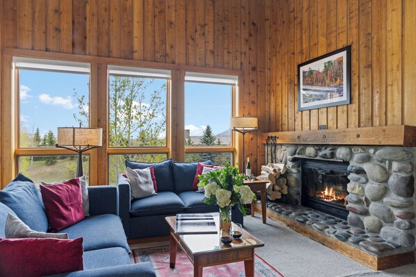 A cozy living room with a stone fireplace, a blue sofa, and a large window offering a scenic view. - Outpost: Nez Perce D5- Hot Tub Access (Teton Village)