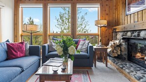 A cozy living room with a sofa, a coffee table with glasses and a vase of flowers, a fireplace, and large windows with a view of trees. - Outpost: Nez Perce D5- Hot Tub Access (Teton Village)
