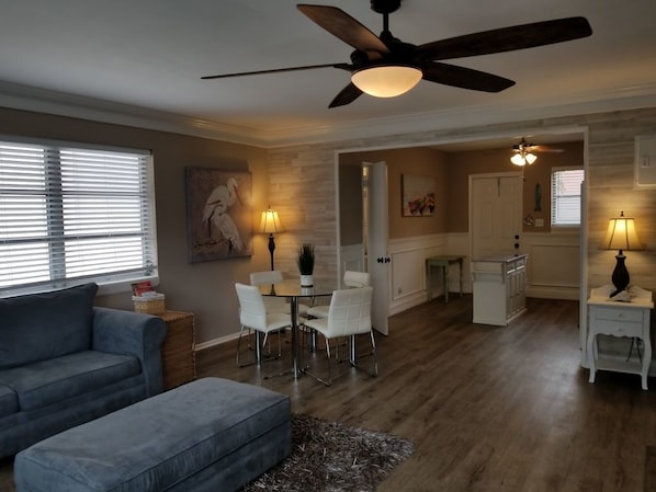 A spacious living area with a ceiling fan, a dining table with chairs, a sofa, and a kitchen in the background.